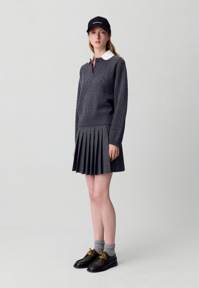 Maglione grigio a maglia grossa con colletto bianco, gonna grigia plissettata, calze grigie e scarpe scure con lacci gialli. Berretto nero indossato.