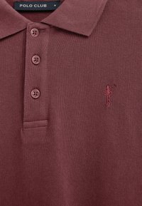 Polo bordeaux avec trois boutons, un petit logo de golfeur brodé sur la poitrine, et une étiquette de col noire indiquant "POLO CLUB" taille M.