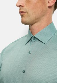 Knopfhemd in Teal mit einem strukturierten geometrischen Muster, ausgestattet mit einem klassischen Kragen und zwei Knöpfen an der Knopfleiste.