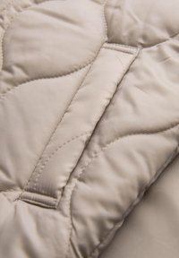 Tessuto beige trapuntato con una superficie liscia e una leggera lucentezza, caratterizzato da un motivo di cucitura ondulato e un pannello inserito per dare struttura.