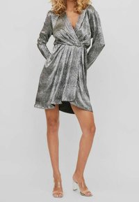 Robe portefeuille en métal argenté avec des manches longues, jupe plissée, décolleté en V profond et ourlet asymétrique. Associée à des sandales à talons transparentes.