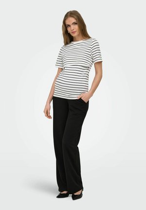 Femme portant un haut à manches courtes rayé blanc et noir, un pantalon noir à jambes larges et des chaussures pointues noires, debout avec une main dans la poche.