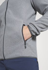 Grå fleecejacka med en zippered ficka och kontrasterande kant. Mjuk textur, figursydd design och en fullständig dragkedjestängning.