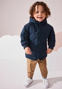 Marinblå quiltad jacka med huva, framzipper och fickor, kombinerad med kaki byxor och vita sneakers med röda detaljer. Ljus bakgrund.