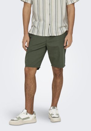 Hombre vistiendo pantalones cortos verdes hasta la rodilla, zapatillas blancas y verdes, y una camisa de manga corta con botones y rayas verticales.