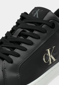 Dettaglio ravvicinato di una scarpa da ginnastica Calvin Klein in pelle nera con lacci neri e logo bianco sul lato, sopra una suola bianca.