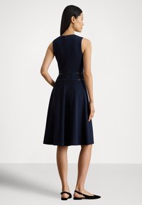 Navyblå ermeløs kjole med tettsittende overdel, utvidet skjørt, glidelås på baksiden og svart dekorativ detalj ved midjen. Semskede slingback-flats.