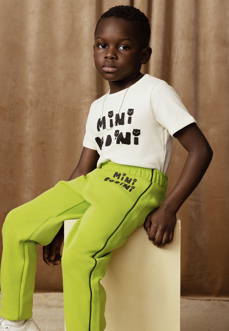 Kind, das ein weißes Baumwoll-T-Shirt mit schwarzem Aufdruck und grüne Sweatpants mit einem Logo trägt, sitzt auf einem hellen Block.