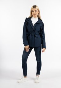 Jachetă impermeabilă de culoare albastru închis, cu glugă cu siret, talie cu curea și buzunare frontale, purtată peste un pulover alb cu guler înalt și blugi negri.