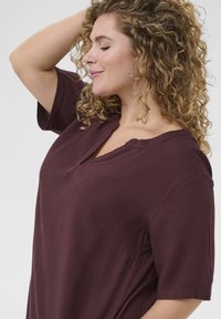 Burgundy blouse with a relaxed fit, short sleeves, notched collar, and soft texture. Design features stitch detailing and side slits.