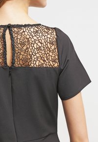 Kvinna som bär en svart klänning med korta ärmar och en genomskinlig blommig spetspanel på den övre ryggen med en liten nyckelhålsöppning och knappstängning.