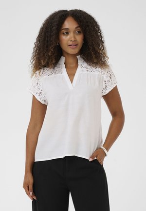 Frau mit lockigem Haar, die eine weiße Bluse mit Spitzenärmeln und schwarze Hose trägt, steht mit einer Hand in der Tasche vor einem einfarbigen Hintergrund.