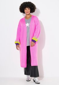 Langer rosa Cardigan mit offenem Front, gerippten Bündchen in Lavendel und Limettengrün. Über einem grauen Shirt mit einem Stern getragen, kombiniert mit schwarzen Hosen und silbernen Stiefeln.