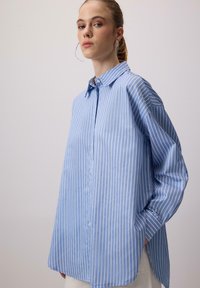 Donna che indossa una camicia button-up a righe verticali blu e bianche, morbida e con colletto, in piedi con una mano in tasca su uno sfondo semplice.