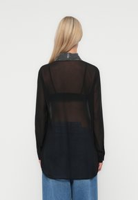Camiseta neagră transparentă cu mâneci lungi, având spatele solid și un guler cu un accent de strasuri. Modelul poartă blugi albaștri cu talie înaltă.