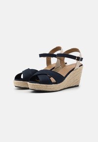 Sandales compensées en tissu bleu marine avec brides croisées, semelle en jute texturée et bride de cheville fermée par une boucle dorée. Doublure intérieure lisse.