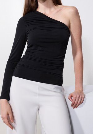 Kobieta ubrana w czarną bluzkę na jedno ramię z długim rękawem, w połączeniu z białymi spodniami z wysokim stanem, stojąca z jedną ręką opartą na powierzchni.