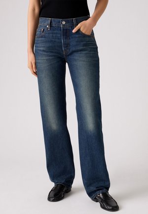 Person trägt dunkelblaue Jeans mit geradem Bein und ausgewaschenen Stellen, schwarze Loafers und ein schwarzes Oberteil, steht mit einer Hand in der Tasche.