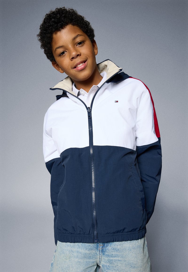 Navy, weiß und rot farblich abgesetzte Windjacke mit Kapuze, Reißverschluss und elastischem Saum; getragen über einem Hemd mit Kragen und Jeans.