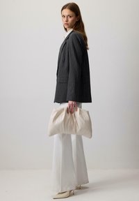 Vrouw in een gestreepte blazer, witte flared broek en hakken, met een grote beige clutch tegen een effen lichte achtergrond.