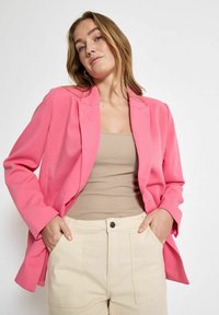 Ryškiai rožinė švarkas su pritaikytu dizainu, su iškarpytomis apykaklėmis ir ilgomis rankovėmis, dėvimas ant smėlio spalvos marškinėlių ir grietinėlės spalvos kelnėmis.