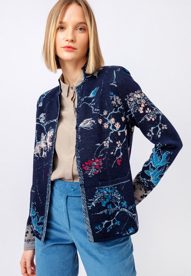 Pletený navy kardigan s květinovými vzory v růžové, modré a červené. Disponuje zapínáním na knoflíky a hladkým povrchem. Sestaveno s lehkou béžovou blůzou.