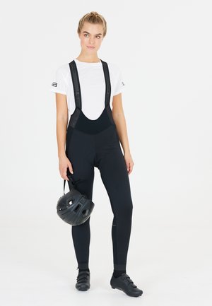 Sorte cykelbukser med mesh-stropper, tætsiddende design, kombineret med en hvid t-shirt og sorte cykelsko. Holder en sort hjelm.