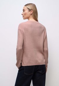 Vrouw die zijwaarts staat in een geribde, stoffige roze trui en donkere jeans tegen een effen lichtgrijze achtergrond.