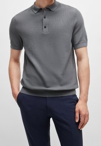 Homme portant un polo en tricot gris à manches courtes avec trois boutons et un pantalon bleu marine, les mains dans les poches, se tenant devant un fond uni.