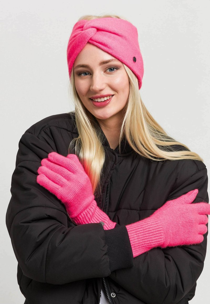 Bandă de cap tricotată roz strălucitor cu un design împletit, asociată cu mănuși lungi roz cu manșete cu arcuri. Modelul poartă o geacă neagră din puf.