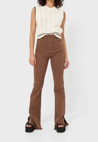 Vrouw in een mouwloze witte gebreide sweater vest, hooggetailleerde bruine flared broek met voren openingen, en zwarte platform sandalen.