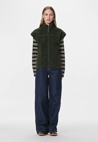 Oļīvzaļš flīsa veste ar augstu apkakli un rāvējslēdzēju, uzvilkta pār melni baltu svītrainu garroku kreklu; kombinēta ar tumši ziliem platajiem džinsiem.