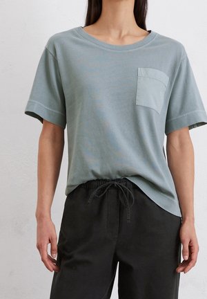 Persoon die een lichtblauw T-shirt met korte mouwen en een borstzakje draagt en zwarte broek met trekkoord, tegen een effen achtergrond.