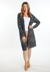 Lange, vorne offene Strickjacke in einem Schwarz-Weiß-Mix, aus strukturiertem Garn gefertigt, kombiniert mit einem weißen T-Shirt und beigen, eng anliegenden Hosen.