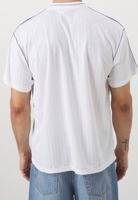 Vit kortärmad skjorta med vertikala ränder, rund krage och lila detaljer på axlarna. Smidig textur, avslappnad passform.