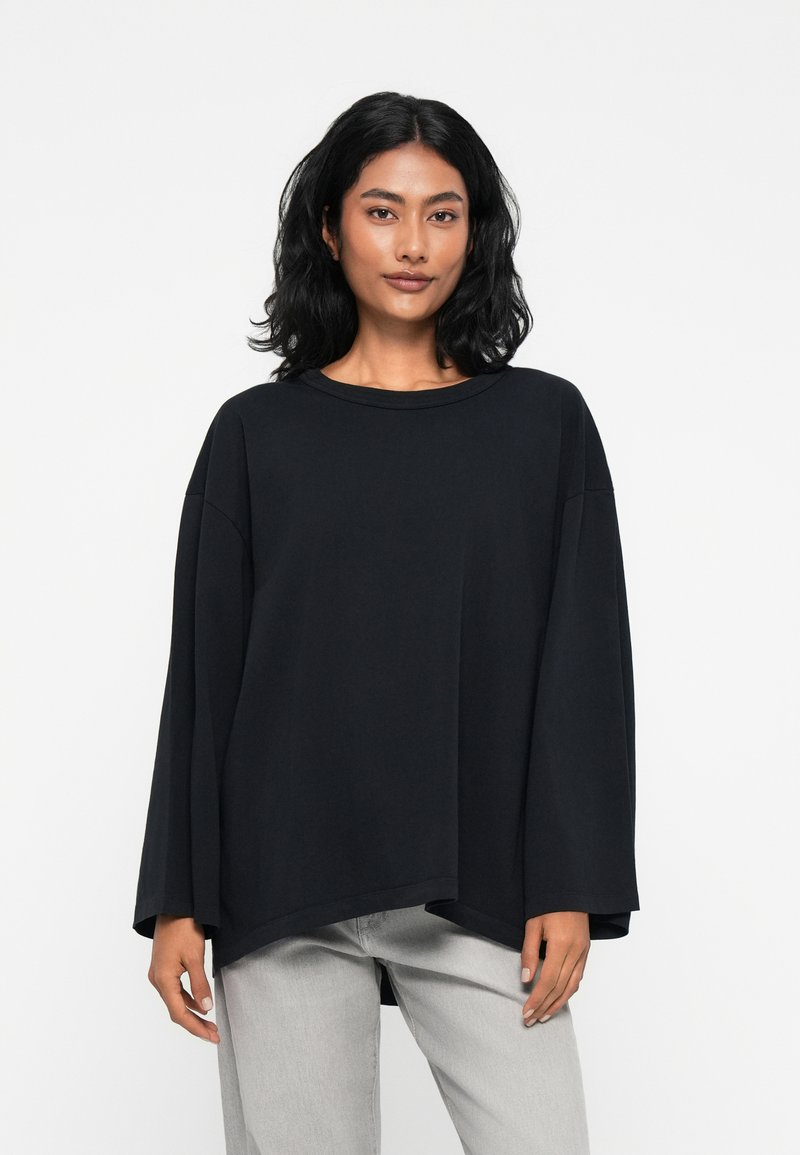 Μαύρη oversized μπλούζα με μακριά μανίκια και στρογγυλή λαιμόκοψη, με χαμηλούς ώμους, φτιαγμένη από μαλακό βαμβακερό ύφασμα, συνδυασμένη με ανοιχτό γκρι τζιν.