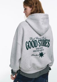 Felpa grigia con cappuccio e grande scritta verde sul retro, con grafica e ricami. Il design è a vestibilità morbida e dalla texture soffice.