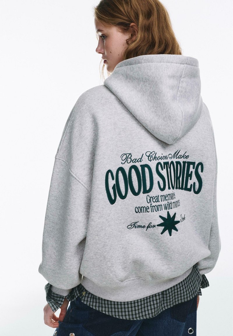 Felpa grigia con cappuccio e grande scritta verde sul retro, con grafica e ricami. Il design è a vestibilità morbida e dalla texture soffice.