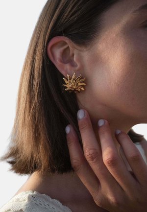 Nahaufnahme einer Frau, die ihr Kinn berührt, mit einem goldenen, sonnenförmigen Ohrring im linken Ohr, kurzen braunen Haaren und hell lackierten Nägeln.