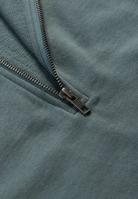 Reißverschlusskleidung aus gedämpftem Teal-Stoff, ausgestattet mit einem Metallreißverschluss und einem sanften Zugtab. Sie verfügt über eine strukturierte, gewebte Innenseite.
