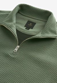Pull-over zippé en molleton vert avec un motif en losanges texturé, fermeture éclair argentée et étiquette en tissu à l'intérieur du col ; taille moyenne.