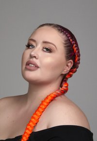 Portrait d'une personne avec une tresse en queue de poisson dégradée du violet à l'orange vibrant, portant un top noir décolleté.