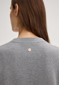 Grijze sweater van een getextureerde stof, met een ronde metalen logo-accent aan de achterkant van de hals. Eenvoudig, klassiek ontwerp.