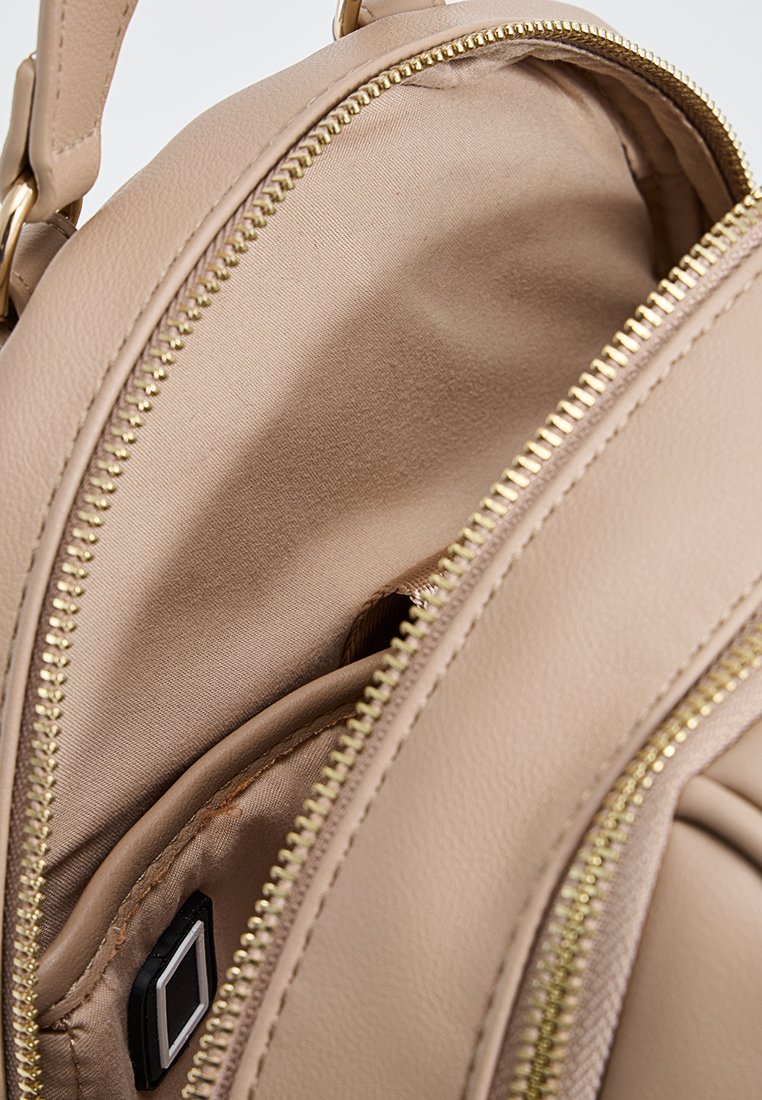 Interno dello zaino in finta pelle beige con una texture liscia, dotato di una cerniera dorata e di una piccola tasca nera per l'organizzazione.