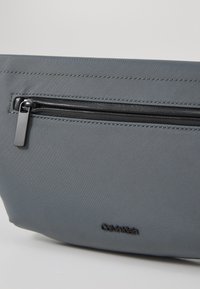 Synteettinen harmaa laukku, jossa on teksturoitu pinta, musta vetoketjupussi ja hienovaraisesti painettu Calvin Klein -logo.