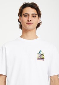 T-shirt blanc à manches courtes avec un graphique coloré d'un chat sur une télévision vintage, portant le texte "VOLCOM". Matière en coton, coupe relax.