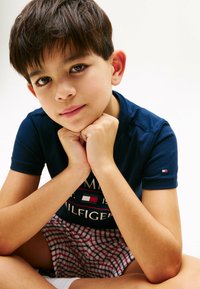 Navyblauwe t-shirt met logo en korte mouwen, gecombineerd met rood-witte geruite shorts, zittende pose, armen onder het kin op de handen gesteund.