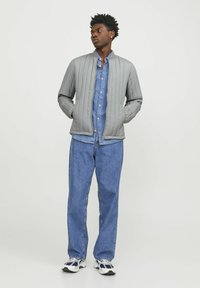 Giubbotto bomber grigio trapuntato con colletto alto, abbinato a una camicia di jeans azzurri e jeans blu larghi, indossato con sneakers bianche e navy.