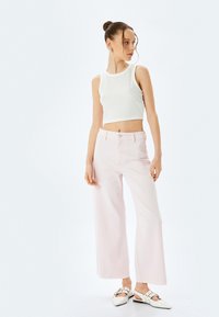 Un crop top blanc à côtes associé à un jean large rose pâle, avec une fermeture à bouton et un ourlet à franges, complété par des chaussures à talons blanches.