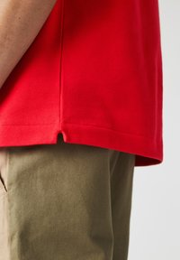 Camisa polo roja hecha de tela texturizada, con una abertura lateral en el dobladillo inferior, combinada con pantalones khaki ligeros.